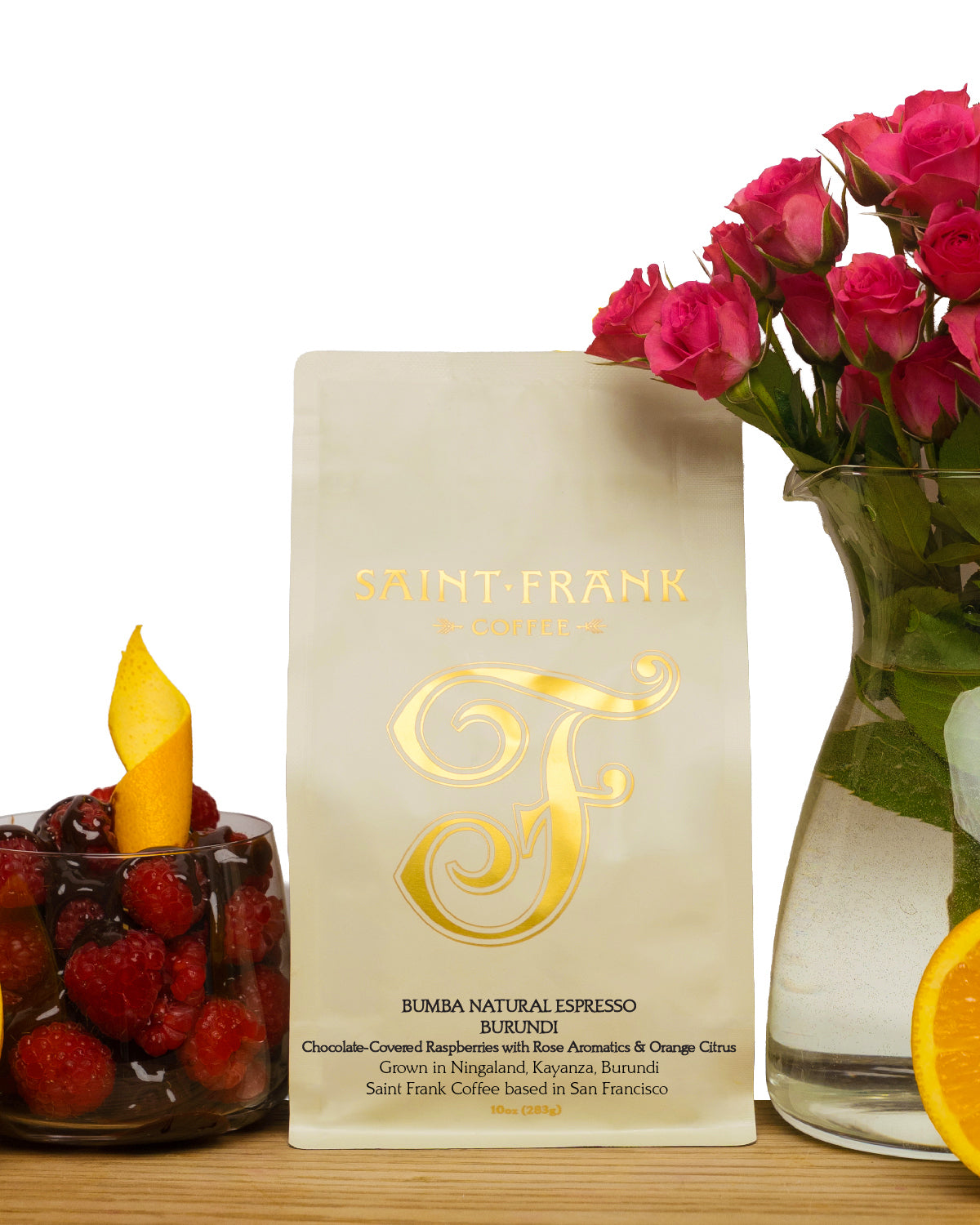 A bag of Saint-Frank Coffee Roasters Rubenci Rumexa Natural Espresso coffee stands on a wooden surface, styled with a vase of pink roses and fruit on a white background.