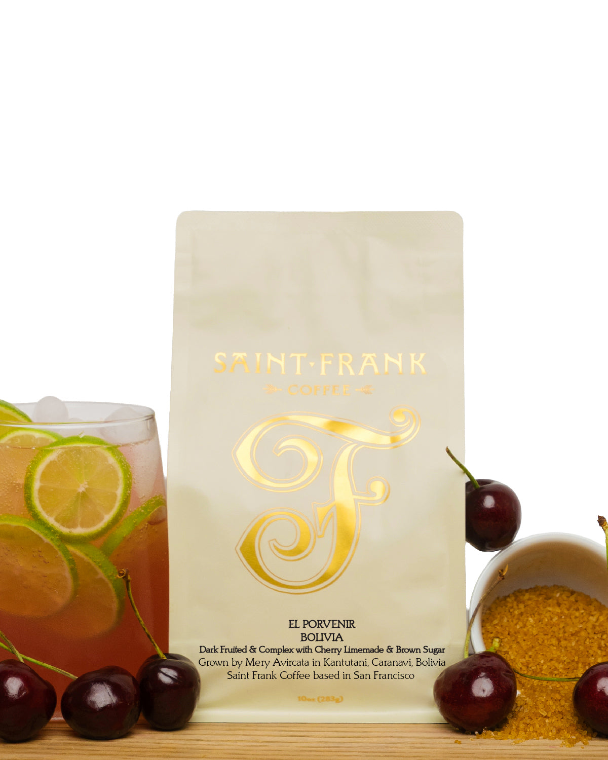 A stand-up pouch of Saint-Frank El Porvenir coffee centered on a white background, styled with cherries, a glass of iced tea with lime, and a bowl of brown sugar on a wooden surface.